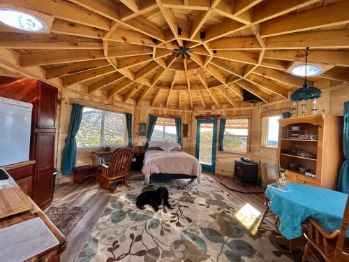 Cozy round cabin interior with wooden beams, a bed, seating area, and a dog on a patterned rug. Large windows offer scenic views.