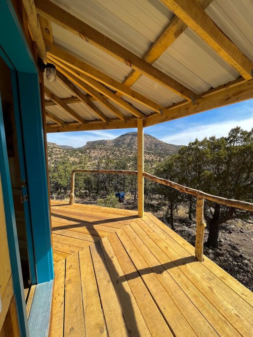 View from a wooden deck with a railing, overlooking a mountainous landscape and clear blue sky.