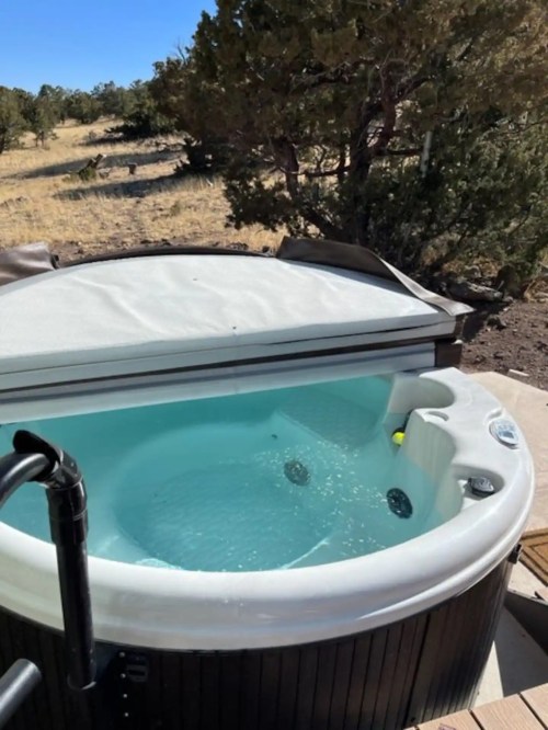 A hot tub with clear water, partially covered, set in a natural outdoor landscape with trees and dry grass.
