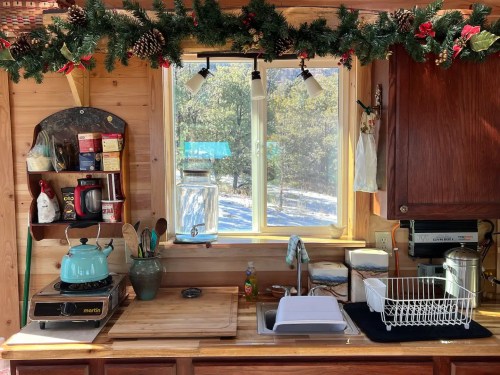 Cozy kitchen with wooden cabinets, a teal kettle, and festive garland above a sunny window overlooking snowy outdoors.