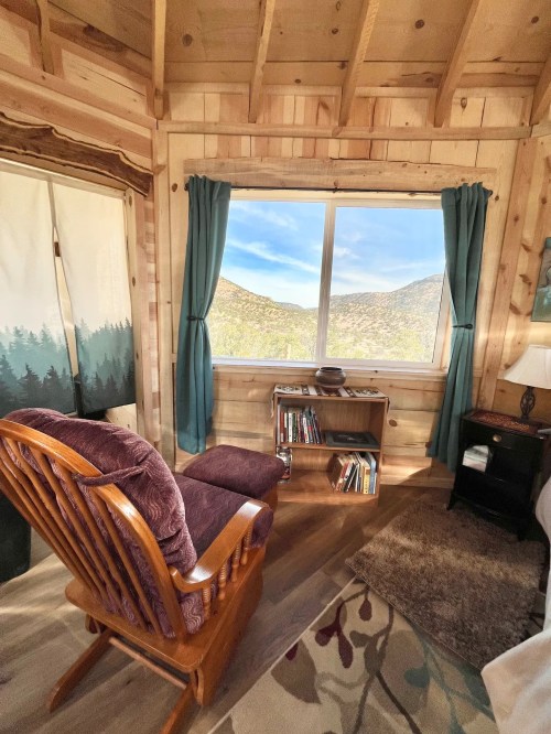Cozy interior of a cabin with a rocking chair, bookshelf, and a large window showcasing mountain views.