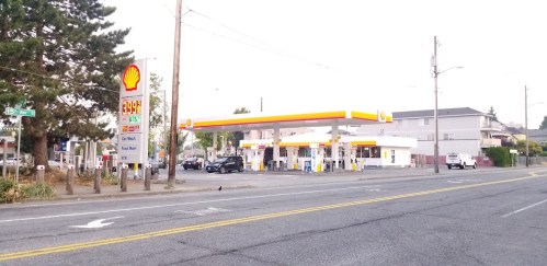 A Shell gas station on a street, featuring fuel pumps and a price sign, with a few cars parked nearby.
