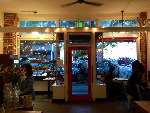 Cozy café interior with large windows, string lights, and patrons enjoying their time, overlooking a street view.