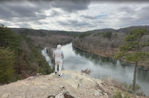 A person in a gray hoodie stands on a rocky ledge overlooking a calm river surrounded by trees under a cloudy sky.