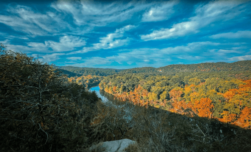 A scenic view of a river winding through colorful autumn trees under a blue sky with fluffy clouds.