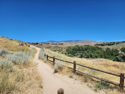 A dirt path winds through dry grass and shrubs, bordered by a wooden fence, with mountains in the background under a clear blue sky.