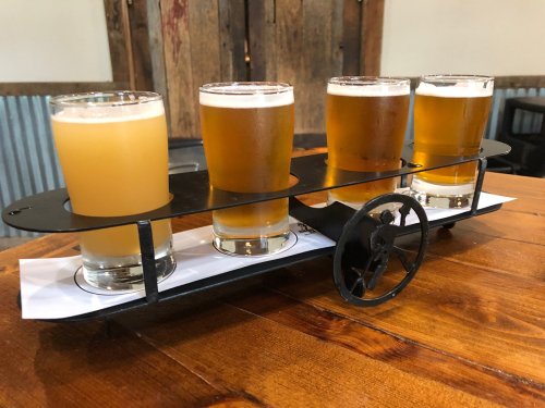 A flight of four beer glasses on a metal tray, set on a wooden table with rustic decor in the background.