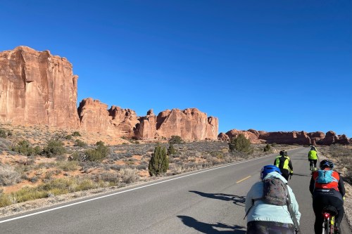 A group of cyclists rides along a road with red rock formations and clear blue skies in the background.