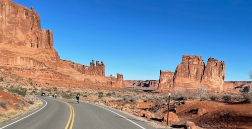 A winding road through red rock formations under a clear blue sky, with cyclists in the distance.