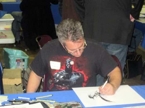 An artist in a Batman t-shirt sketches on paper at a convention, focused on his work.