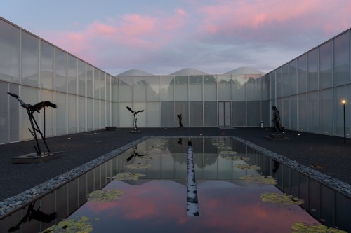 A serene courtyard with sculptures, a reflective pond, and a colorful sky at dusk.