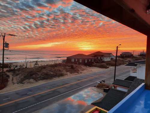 A vibrant sunset over the ocean, with colorful clouds and silhouetted houses along a quiet road.