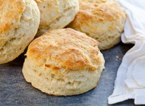Freshly baked, golden-brown biscuits stacked on a dark surface, with a white cloth nearby.