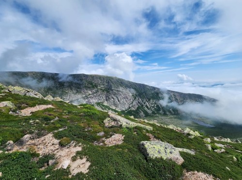 A panoramic view of a mountainous landscape with rocky terrain, lush greenery, and clouds partially obscuring the peaks.
