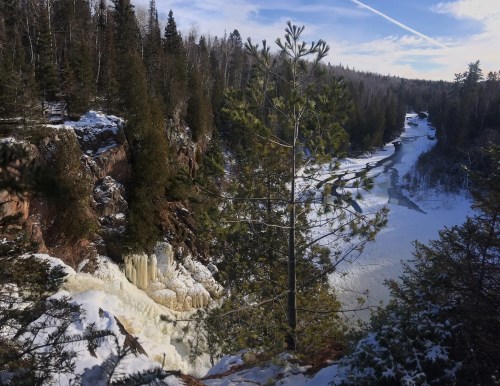 A winter landscape featuring a frozen waterfall, snow-covered trees, and a river winding through a forested area.