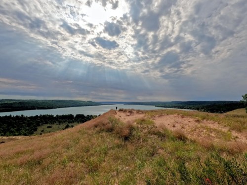 A scenic view of a river surrounded by hills, with dramatic clouds and sunlight breaking through the sky.