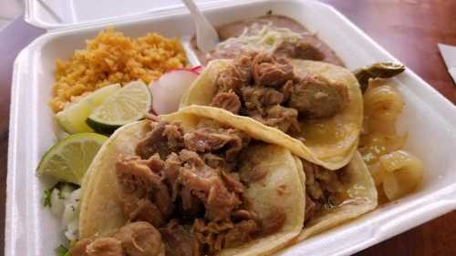 A takeout container with three tacos filled with meat, rice, beans, and garnishes like lime and radishes.