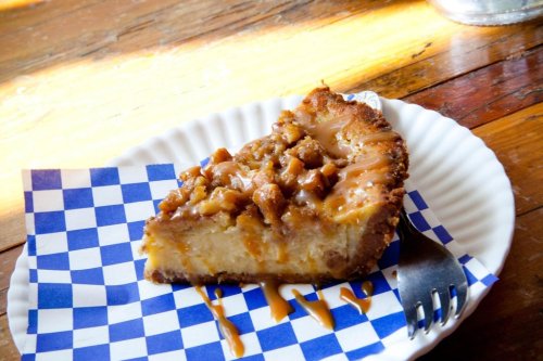 A slice of cheesecake topped with caramel and nuts on a blue checkered paper plate, resting on a wooden table.