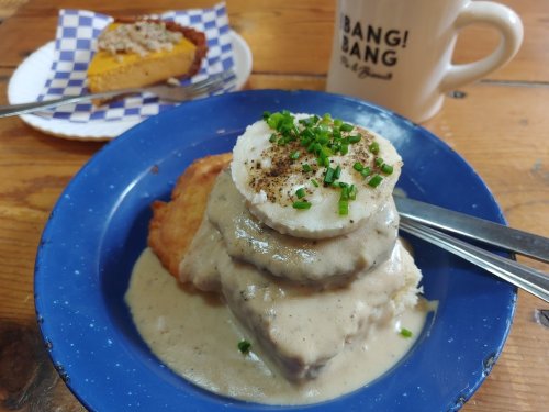 A blue plate with biscuits and gravy topped with a fried egg and chives, alongside a slice of pumpkin pie and a mug.