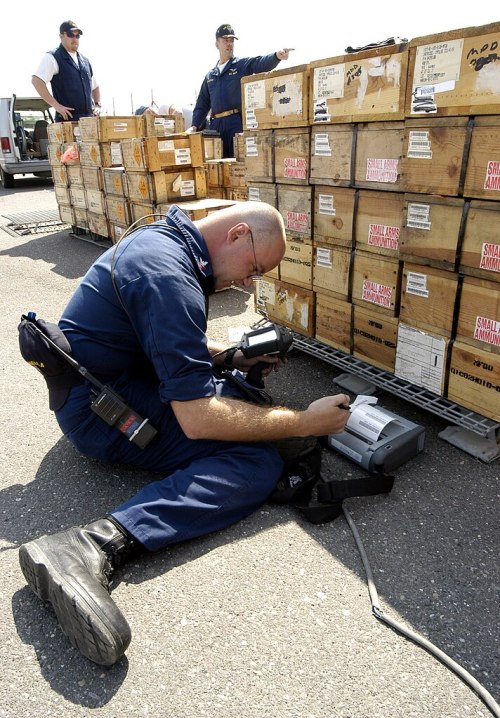 A man in a blue uniform kneels, inspecting crates while using a handheld device, with others in the background.