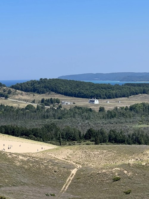 A scenic view of rolling hills, a sandy area, and a distant blue lake under a clear sky.