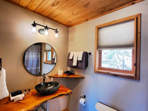 A modern bathroom featuring a round mirror, wooden shelves, a stone sink, and natural light from a window.