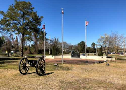 A cannon in a park with flags and a memorial stone surrounded by trees and grass under a clear blue sky.