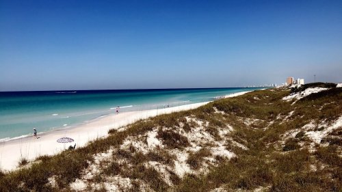 A serene beach scene with white sand, gentle waves, and a clear blue sky, featuring distant buildings along the shoreline.