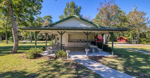 A picnic shelter with tables surrounded by trees, with a red building visible in the background on a sunny day.