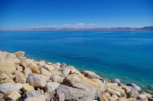 A serene view of calm blue water meeting a rocky shoreline under a clear blue sky. Mountains are visible in the distance.