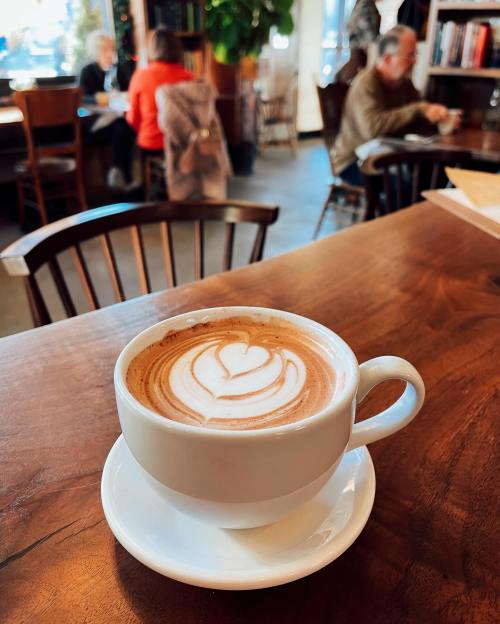 A latte with a heart design sits on a wooden table in a cozy café, with people chatting in the background.