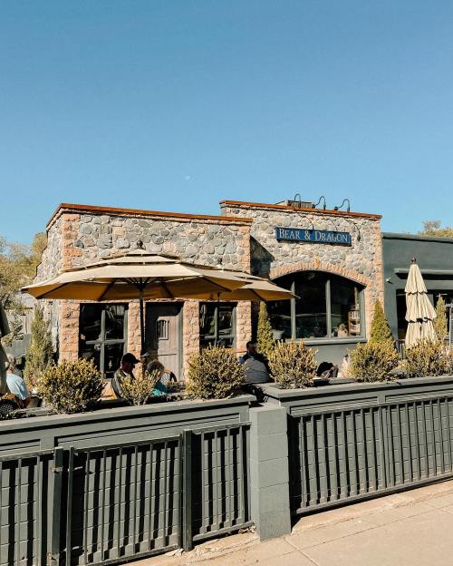 A stone building with a sign reading "Bear & Dragon," featuring outdoor seating and umbrellas under a clear blue sky.