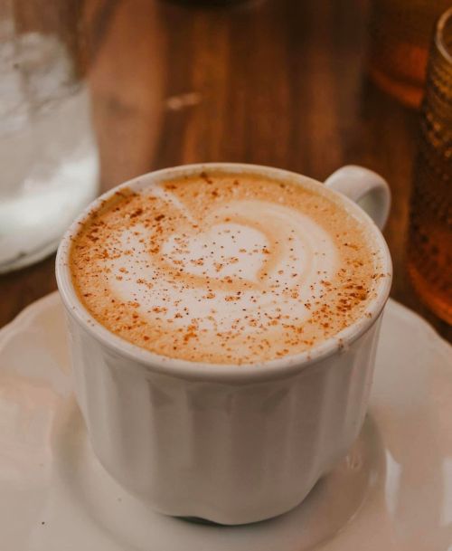 A white cup of coffee with a heart-shaped foam design, sprinkled with cinnamon, on a wooden table.