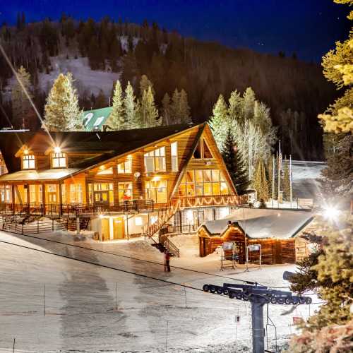 A cozy ski lodge illuminated at night, surrounded by snow and pine trees, with a ski lift in the foreground.