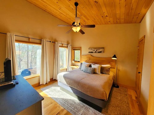 Cozy bedroom with wooden ceiling, large windows, a king-sized bed, and a desk in natural light.
