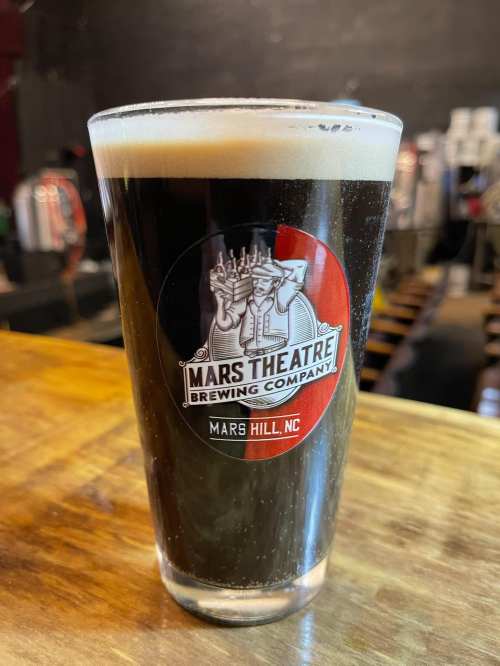 A pint glass of dark beer with a logo for Mars Theatre Brewing Company, set on a wooden table.