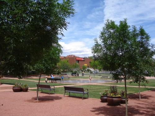 A park scene with green trees, benches, and a water feature, under a blue sky with scattered clouds.