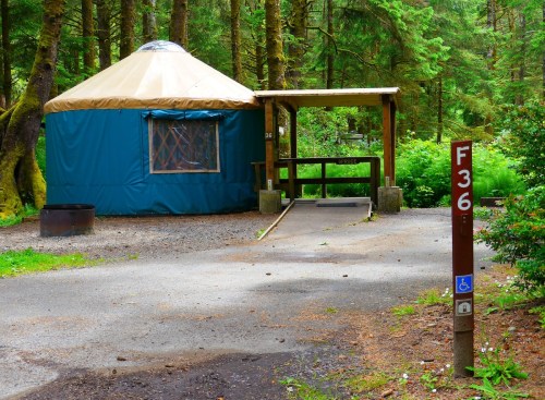 A blue yurt in a forested area, with a wooden porch and a sign indicating a nearby accessible campsite.