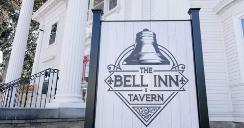 Sign for "The Bell Inn & Tavern" featuring a bell graphic, in front of a large, white-columned building.