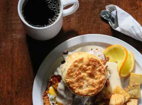 A plate with a biscuit sandwich, gravy, potatoes, and orange slices, alongside a cup of black coffee.