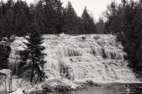 A black and white image of a cascading waterfall surrounded by snow-covered trees and rocky terrain.