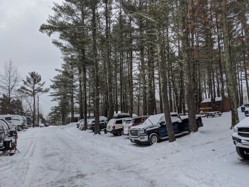 Snow-covered parking area surrounded by tall trees, with several vehicles lined up along a snowy road.