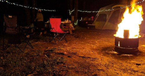 A cozy campsite at night with a roaring fire, two chairs, and string lights in the background.
