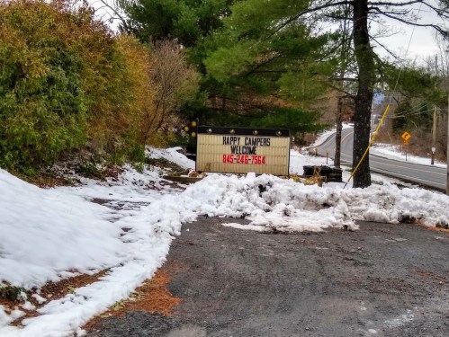 Sign reading "Happy Campers Welcome" at a snowy roadside, with trees and a partially cleared path.
