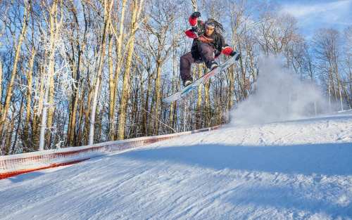 A snowboarder jumps off a snowy slope, surrounded by trees, with snow spraying up in the air.