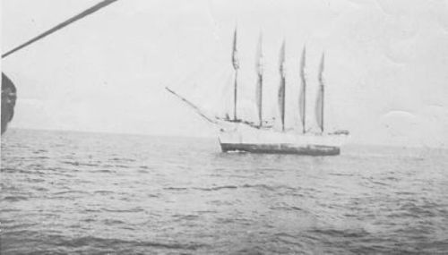 A historic black-and-white photo of a tall ship with five sails sailing on calm waters.