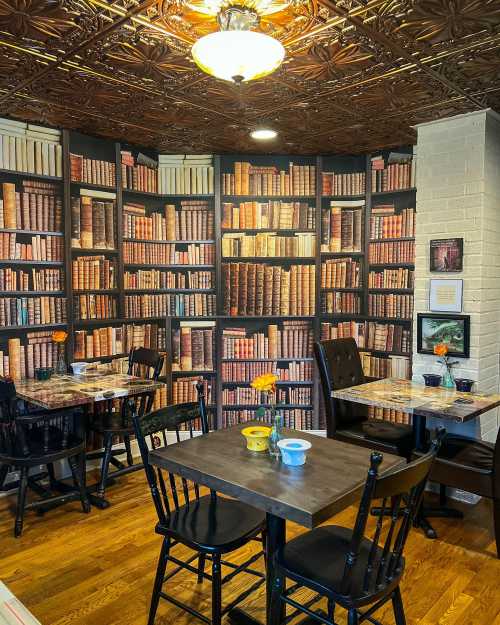 Cozy café corner with wooden tables, black chairs, and a wallpaper of bookshelves, featuring a flower on the table.