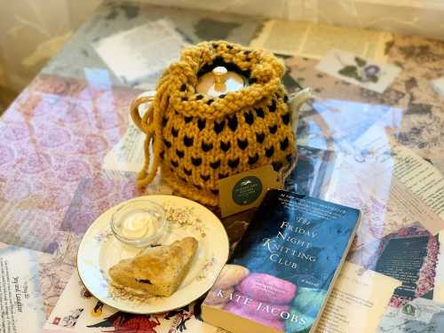 A cozy scene featuring a knitted teapot cozy, a slice of cake on a plate, and a book titled "The Friday Night Knitting Club."