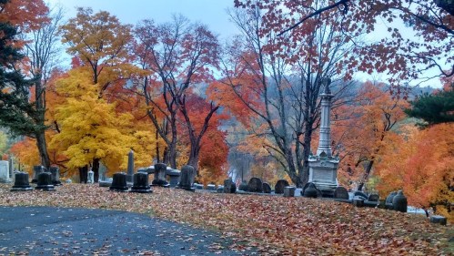 A serene cemetery scene with colorful autumn trees and fallen leaves, featuring gravestones and a tall monument.