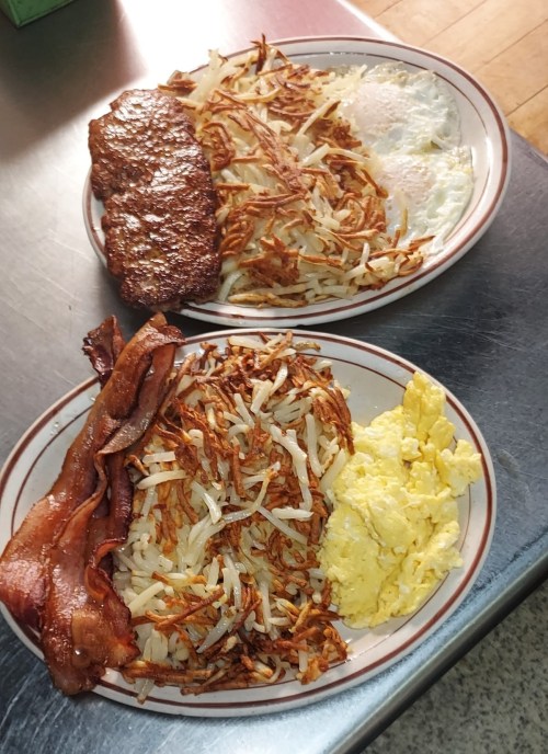 Two plates of breakfast: one with hash browns, eggs, and bacon; the other with hash browns and a beef patty.